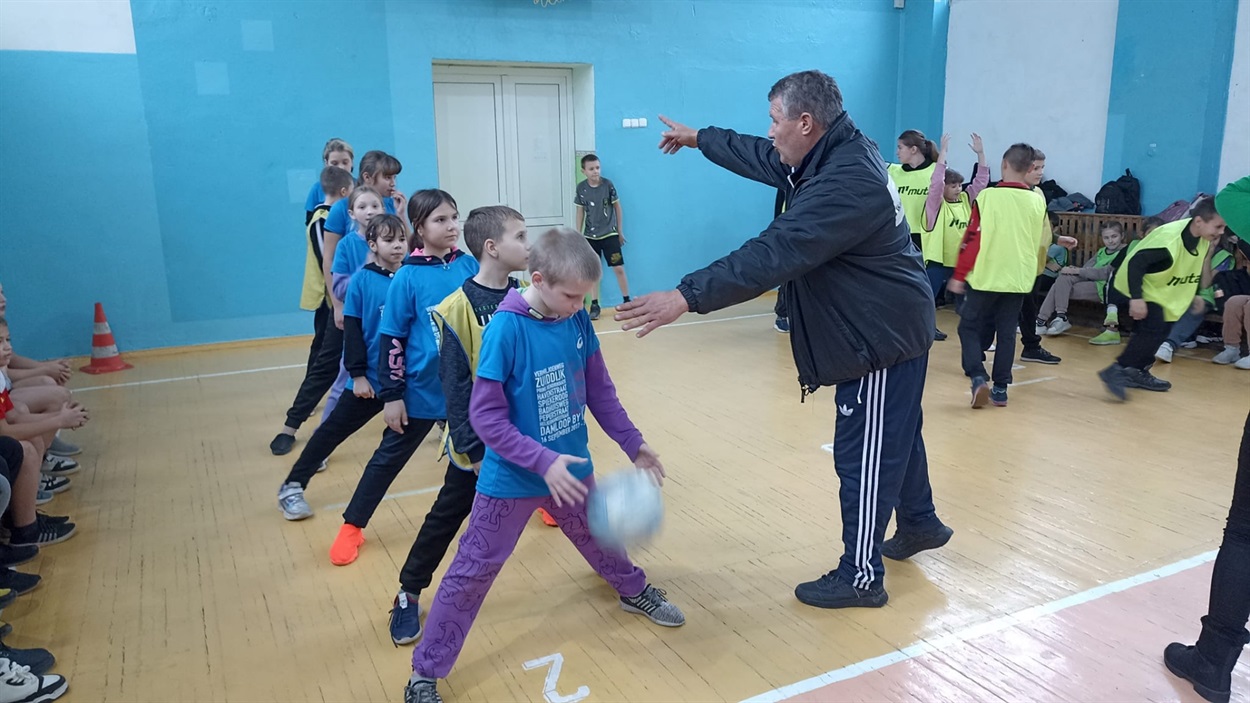 Командам пояснюють як виконувати активність "Конвеєр" – вправа з мʼячем. Ставши в колону треба мʼяч від першого до останнього члена команди передати над головами, а назад повернути, прокочуючи між ногами